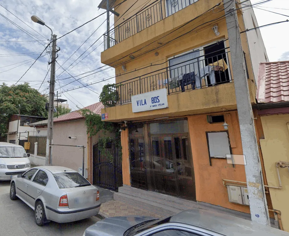Three-story yellow building with a balcony and a sign reading Vila Bos on a sunny street with parked cars nearby.